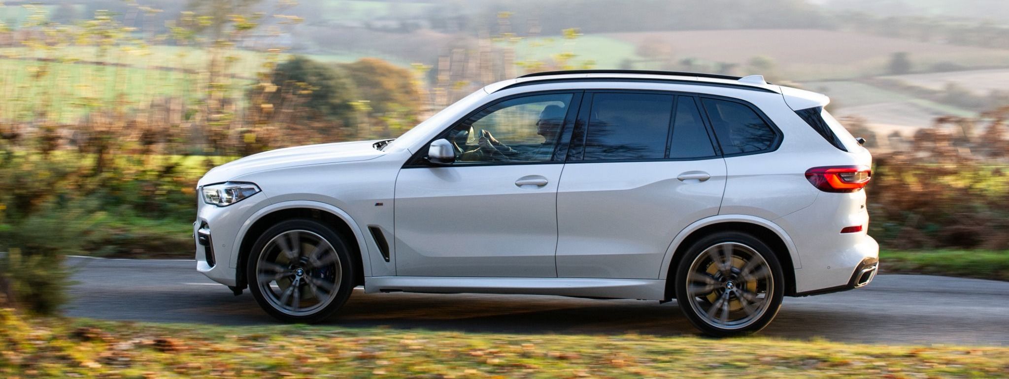 side view white bmw x5 driving down a country road