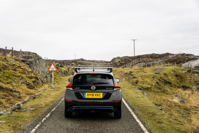 Rear view of a nissan leaf driving on a road surround by fields and with a warning sign next to the car