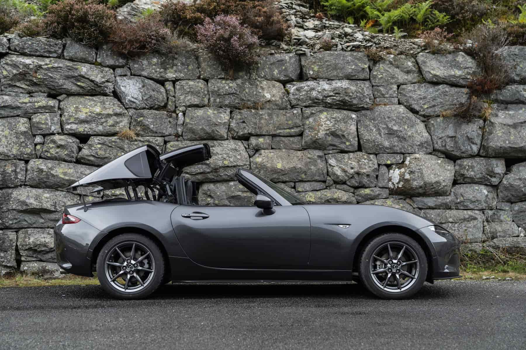 grey mazda mx 5 parked beside a stone wall with the convertible roof coming down