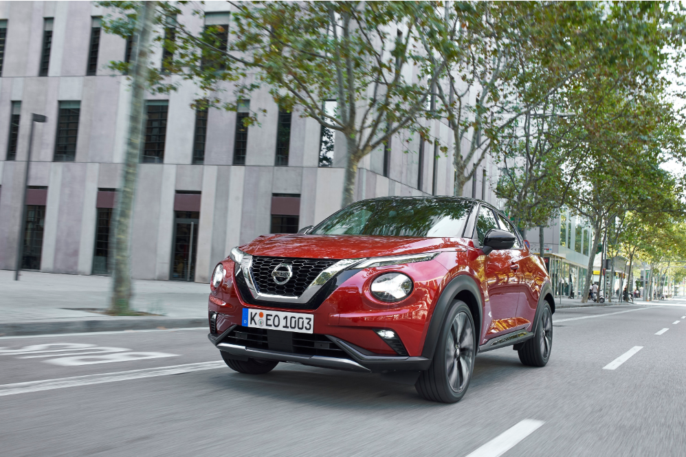front view of a red nissan juke driving down the road with buildings and trees in the background