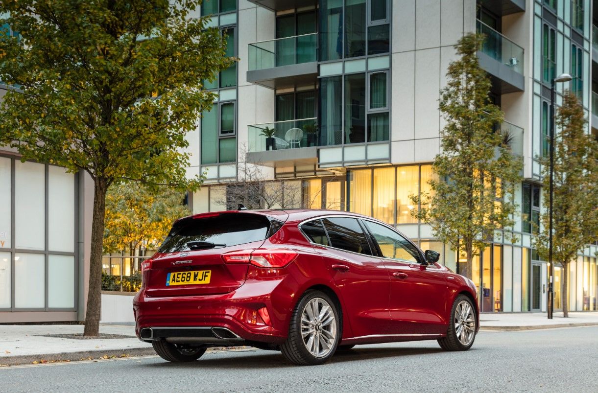side view of a red Ford Focus Vignale parked on the side of a road in front of buildings and trees