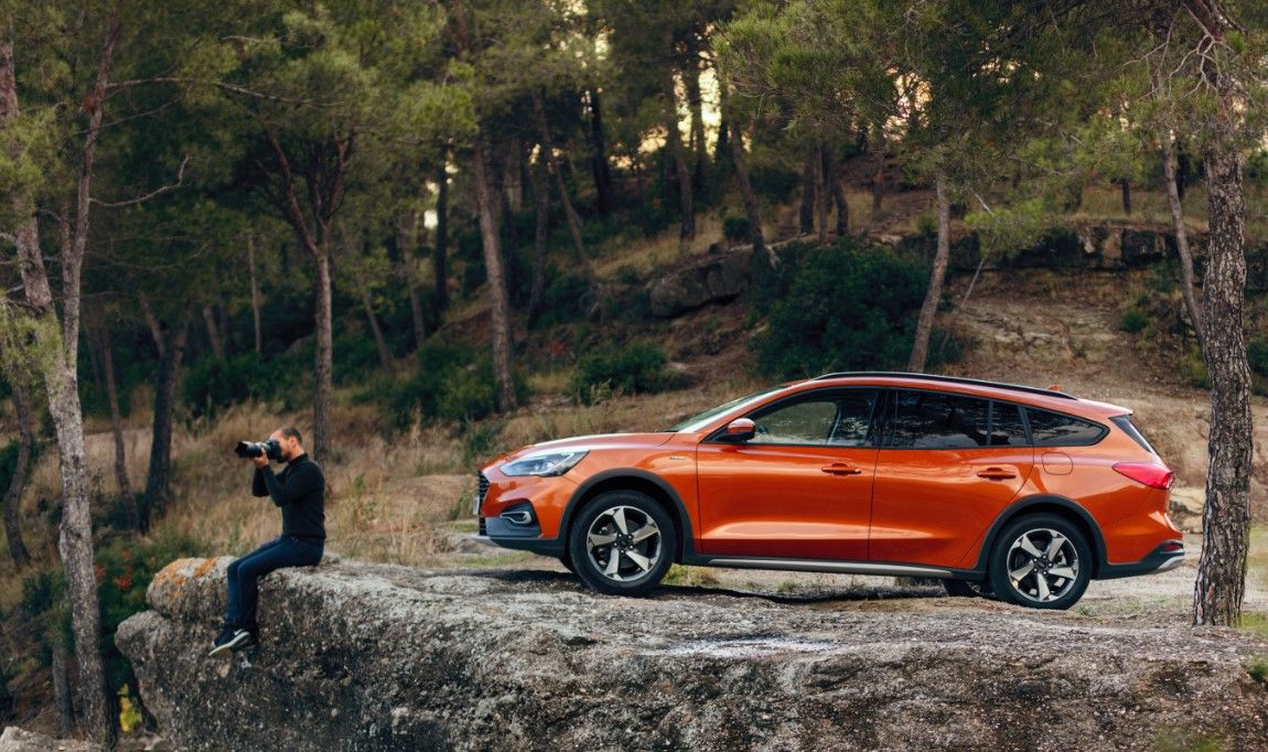 Side view of an orange Ford Focus parked on a cliff with a man sitting on the ledge taking a photo in front of it and trees in the background.