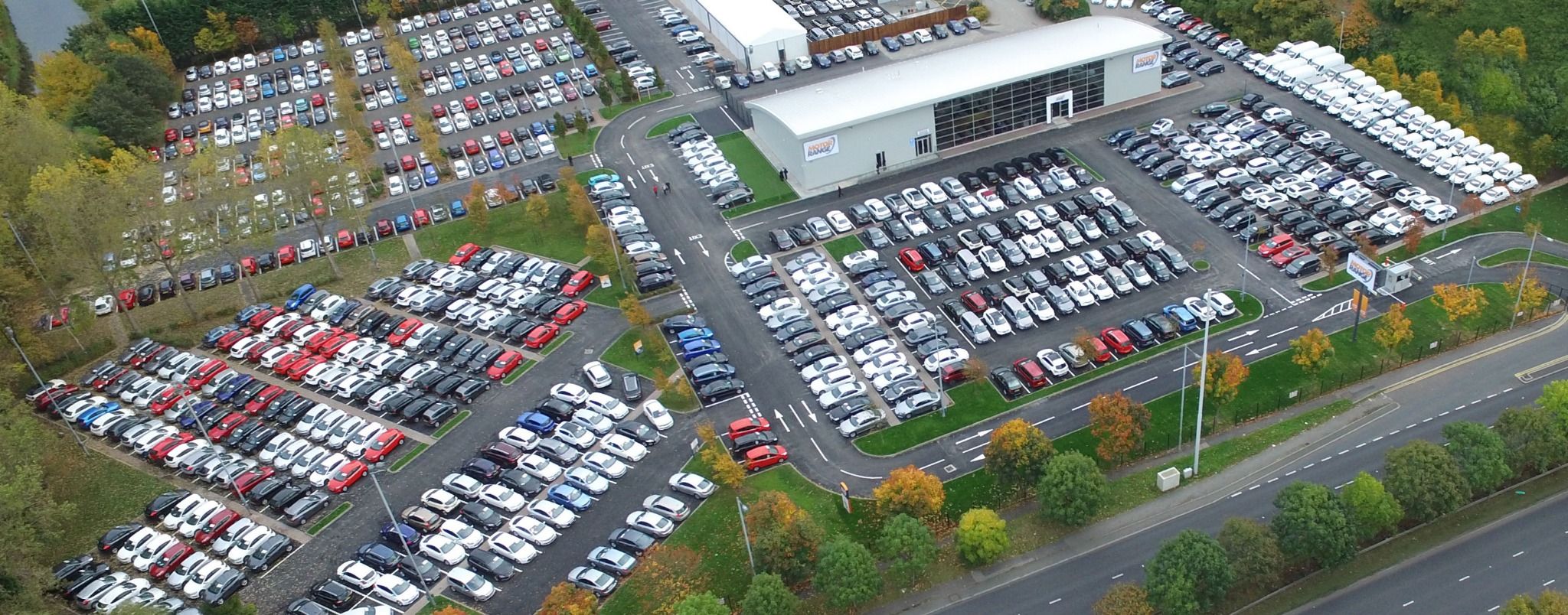 High-angle view of a vast outdoor car lot filled with rows of new and used vehicles of various colors. A large dealership building is visible in the upper center of the image, surrounded by organized parking and access roads. Trees with autumn foliage bor