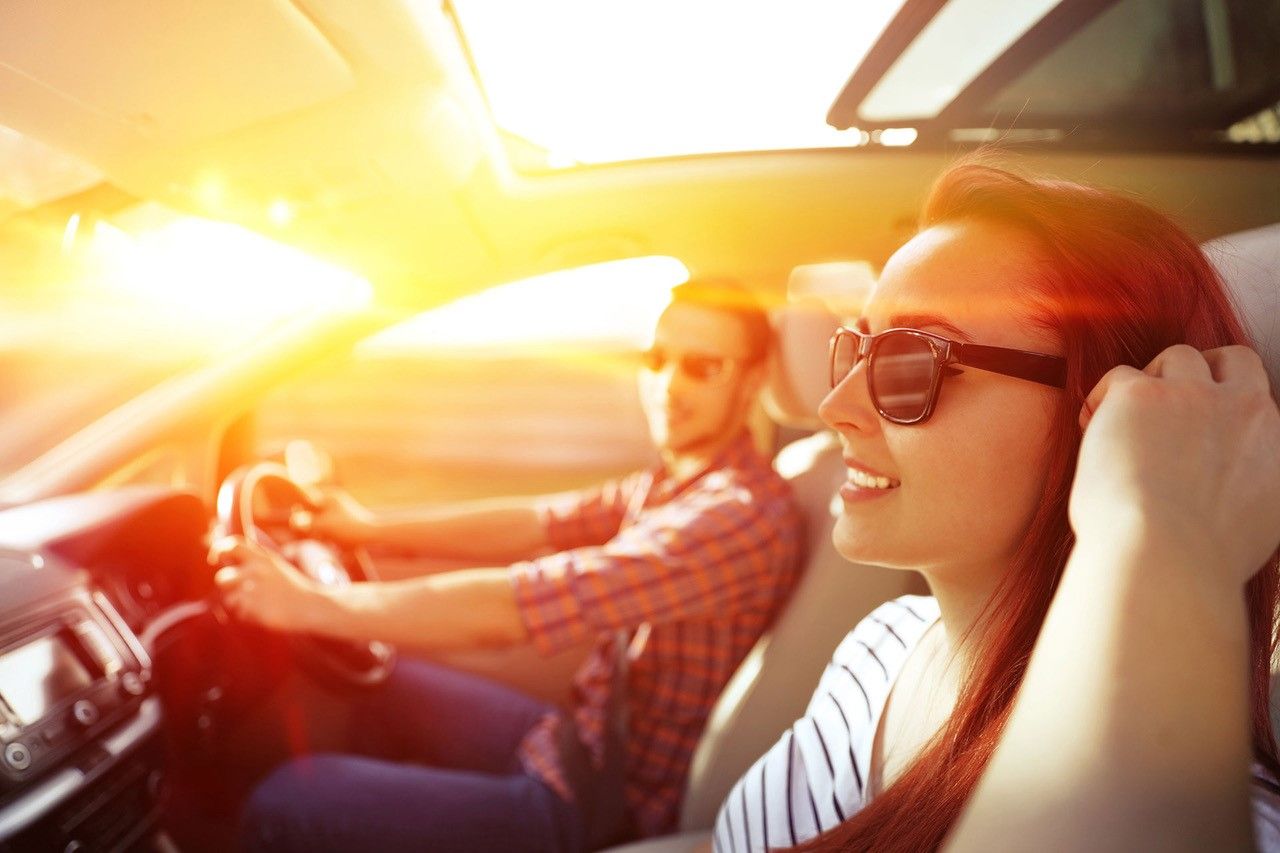 A woman and a man wearing sunglasses sit in a car with sunlight streaming through the windows, creating a warm glow inside.