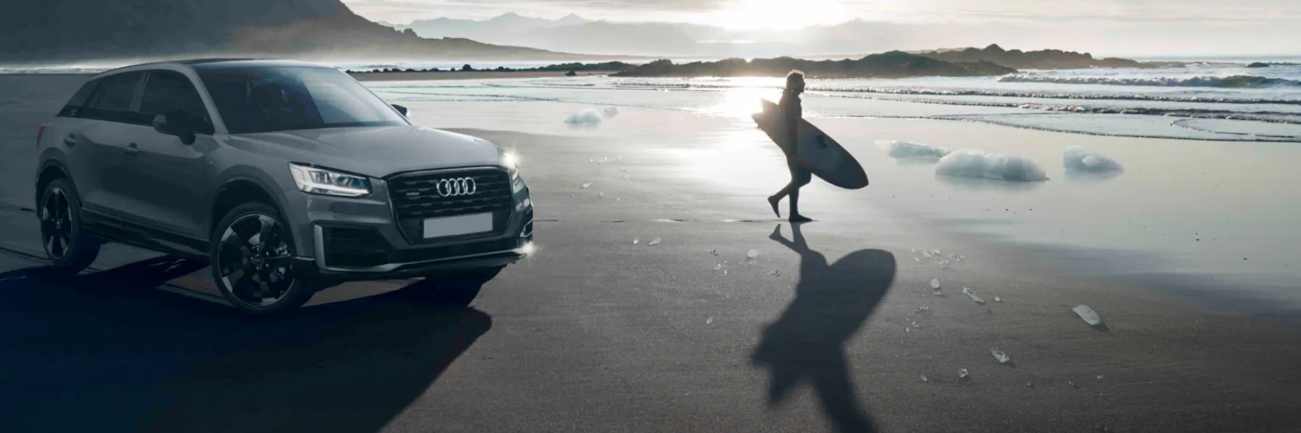 audi on beach with man walking into sea with surfboard