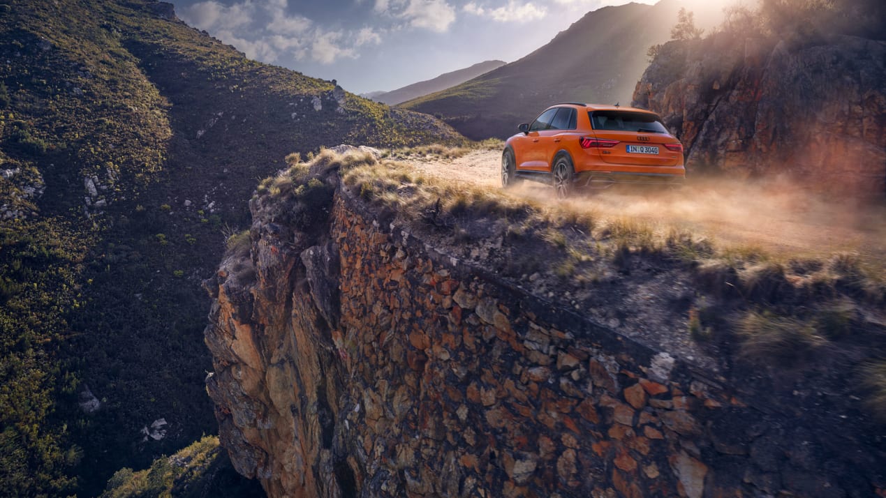 rear side exterior view of an orange audi q3 rear driving down a dirt road with dust billowing behind the car