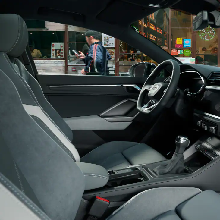 interior view of a left hand drive audi q3 sportback from passenger's side parked up in a busy street. There is a man in the background wearing a sports jacket and on his phone.