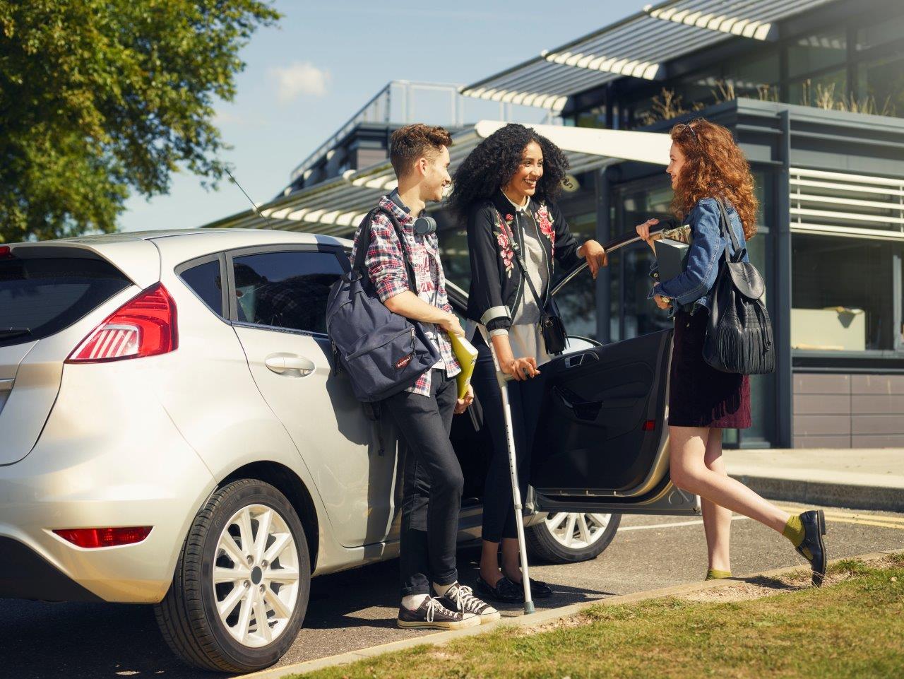 3 young adults standing next to a ford fiesta with one holding a crutch