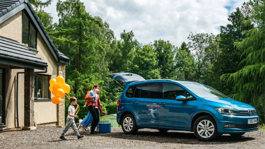 Blue Volkswagen Touran side parked and being loaded with luggage by family