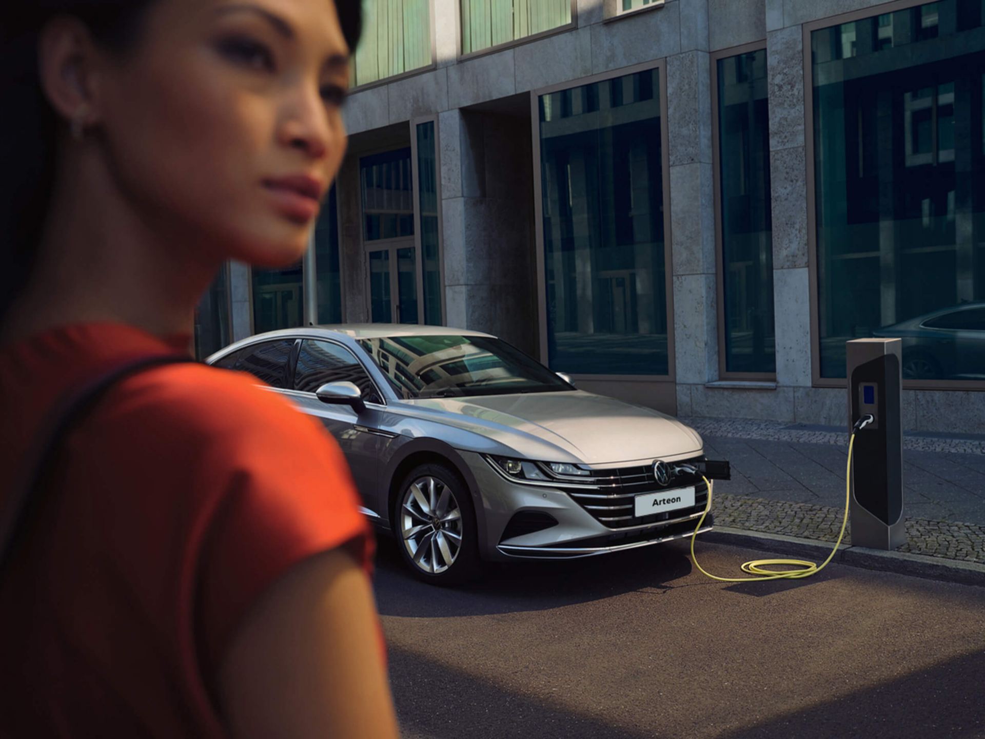 image of a lady wearing orange stood in front of a silver electric volkswagen arteon charging  up in the street